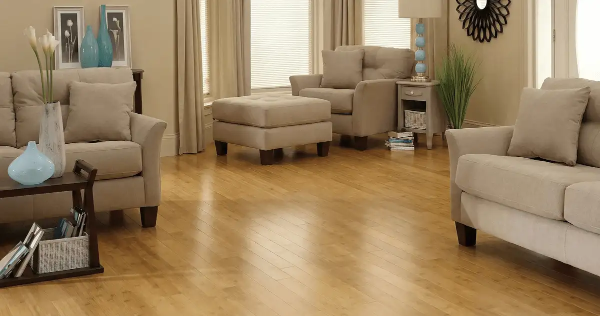 A living room with professionally installed bamboo flooring, beige sofas, a matching ottoman, a side table with a blue lamp, vases, framed pictures, and large windows with blinds and curtains in Ann Arbor, MI